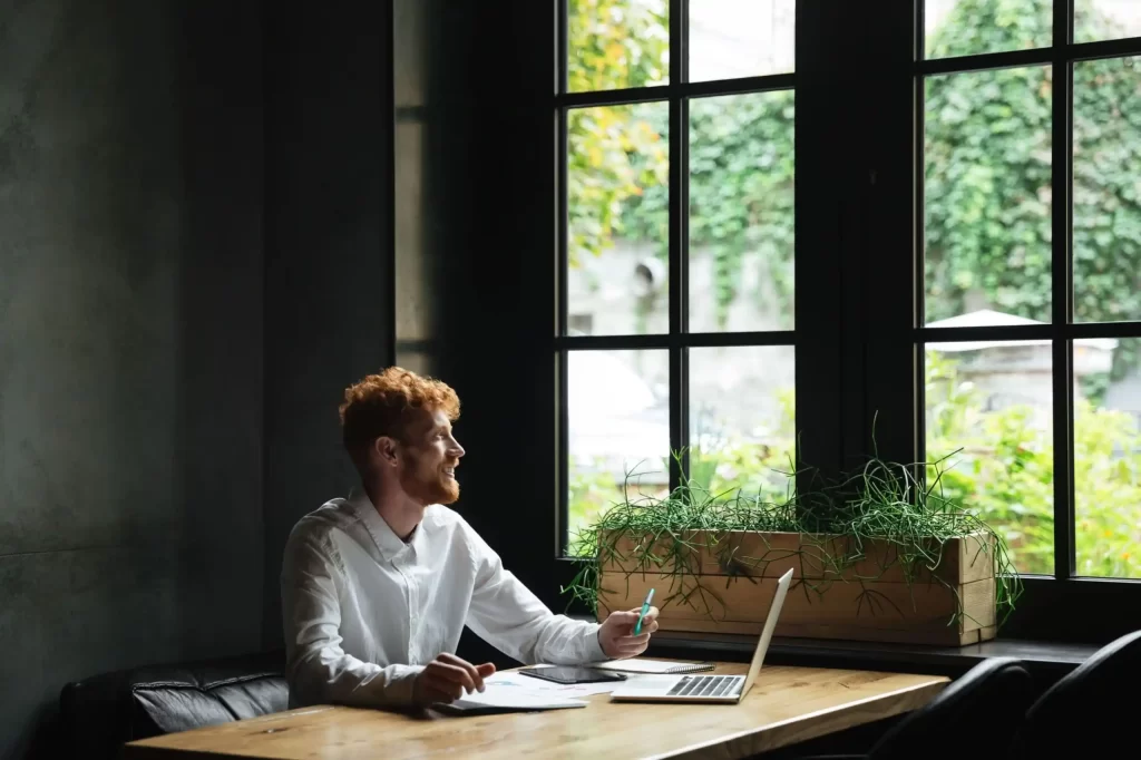 man zit achter zijn laptop en kijkt naar buiten via het raam