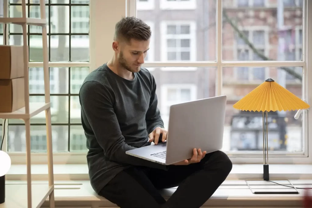man zit op vensterbank bij het raam met laptop op zijn schoot